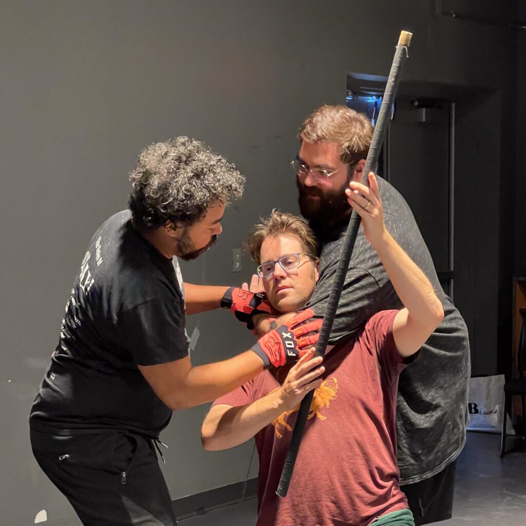 Three men rehearse a staged fight scene indoors. One man wearing glasses and a maroon shirt kneels while holding a long staff across his shoulder. Another man behind him wraps an arm around his neck in a mock chokehold, while a third man in a black shirt and red gloves holds the staff and gestures as if directing or adjusting the action.