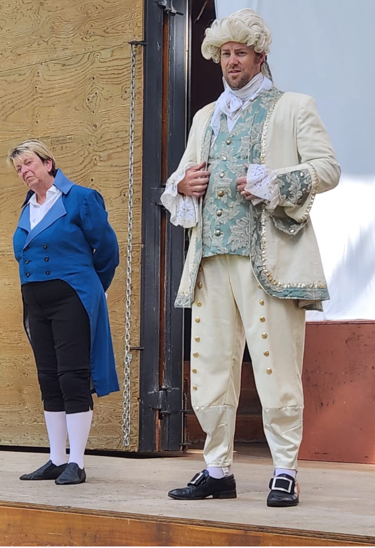 Two performers stand on an outdoor wooden stage in period costumes. On the right, a man wears an elaborate 18th-century style cream suit with gold buttons, a brocade vest, lace cuffs, and a white powdered wig. He looks forward with a slight smile, holding his coat open. On the left, another performer wears a blue tailcoat, white shirt, black breeches, white stockings, and black shoes, standing with hands behind their back and head tilted to the side.