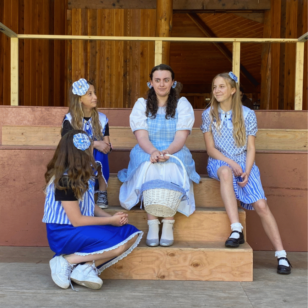 Four young performers in costume sit on wooden steps inside a rustic wooden stage structure. At the center is a person dressed as Dorothy from The Wizard of Oz, wearing a blue gingham dress, white blouse, silver shoes, and holding a white basket. Three girls dressed in blue and white costumes with matching bows and hair accessories sit or kneel around her, smiling and looking at each other.