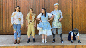 Five actors in costume standing on an outdoor stage. They are dressed as the Scarecrow, the lion Dorothy, the Tin Woodman and Toto from the Wizard of Oz.