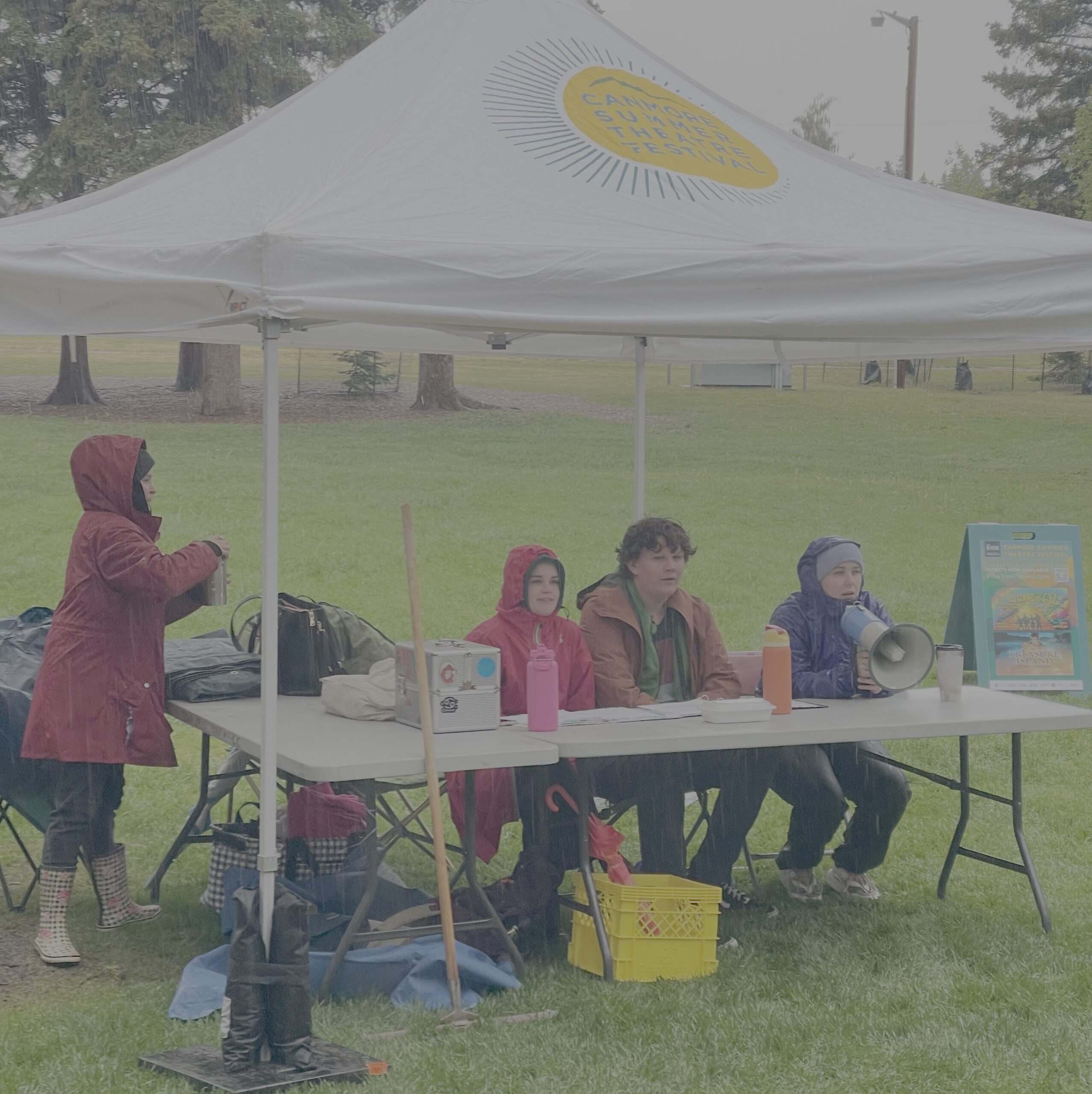 Three people dressed in rain coats sit at a table under at canopy tent during a cold and rainy rehearsal.